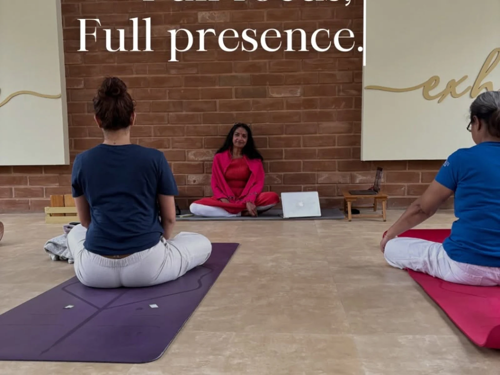 A yoga instructor in a pink outfit sits cross-legged facing participants seated on yoga mats in a brick-walled studio. Text overlay reads, “Full house, Full focus, Full presence.”