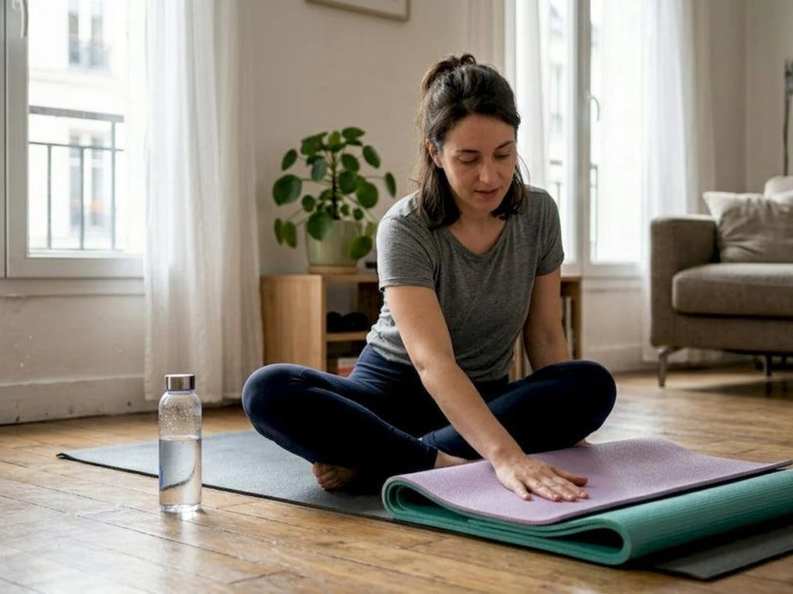A woman sitting cross-legged on a yoga mat in a bright living room, preparing her mat for a home yoga practice.